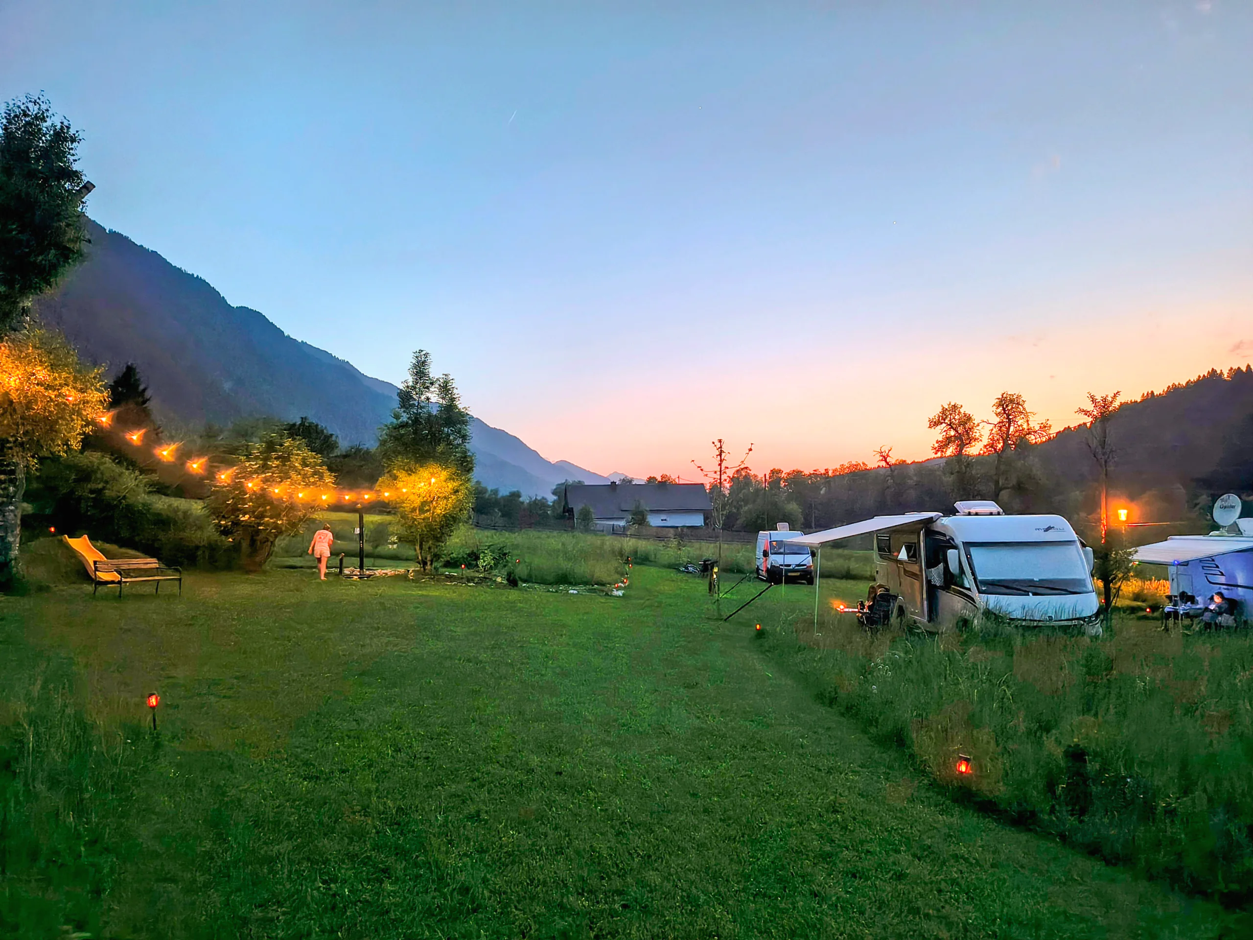 Wohnmobile auf der Wiese neben dem Haus im Abendlicht, dahinter ein bewaldeter Berghang