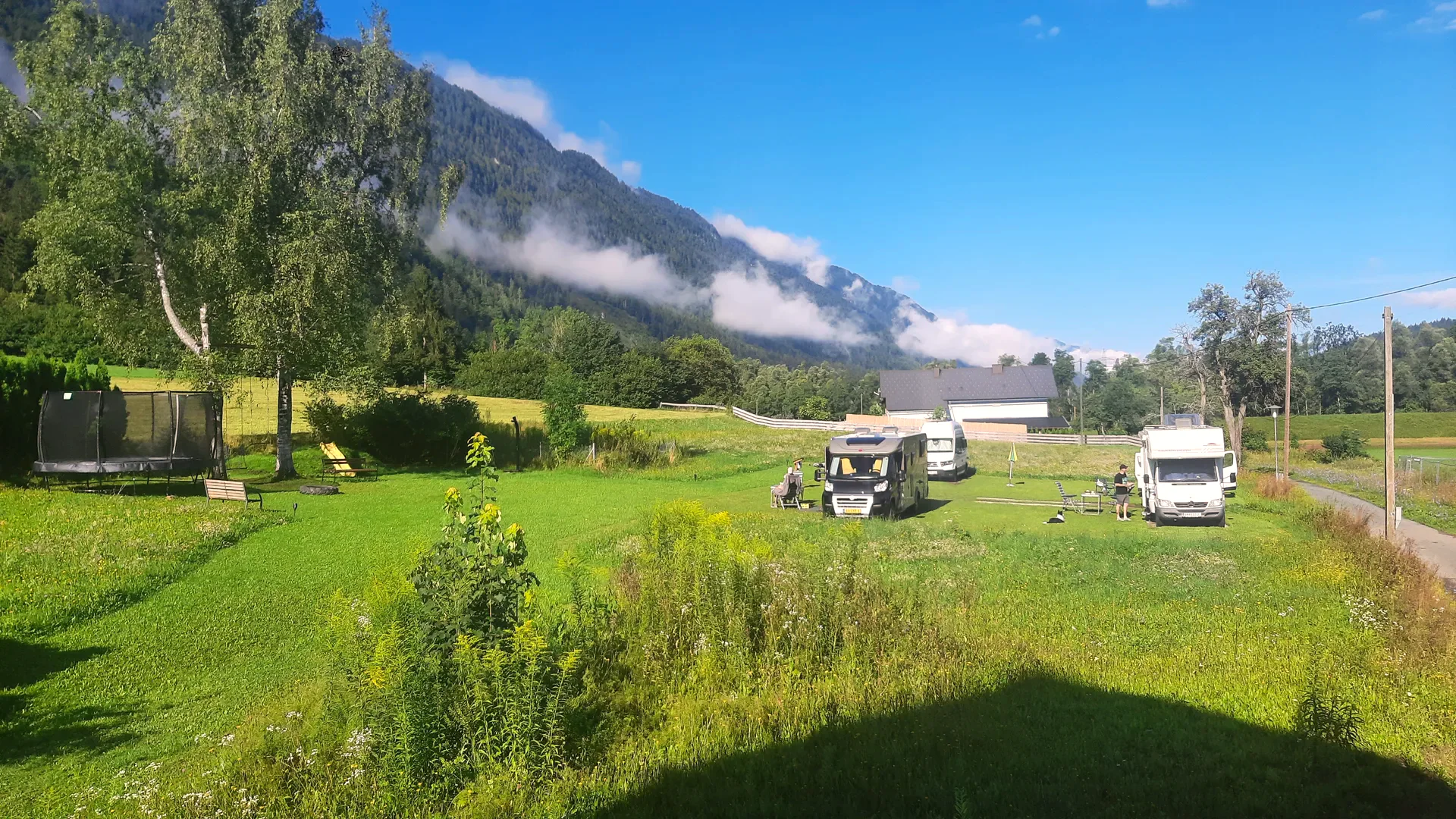 Zwei Wohnmobile auf der Wiese, links das Trampolin, im Hintergrund der Berghang mit tief hängenden Wolken