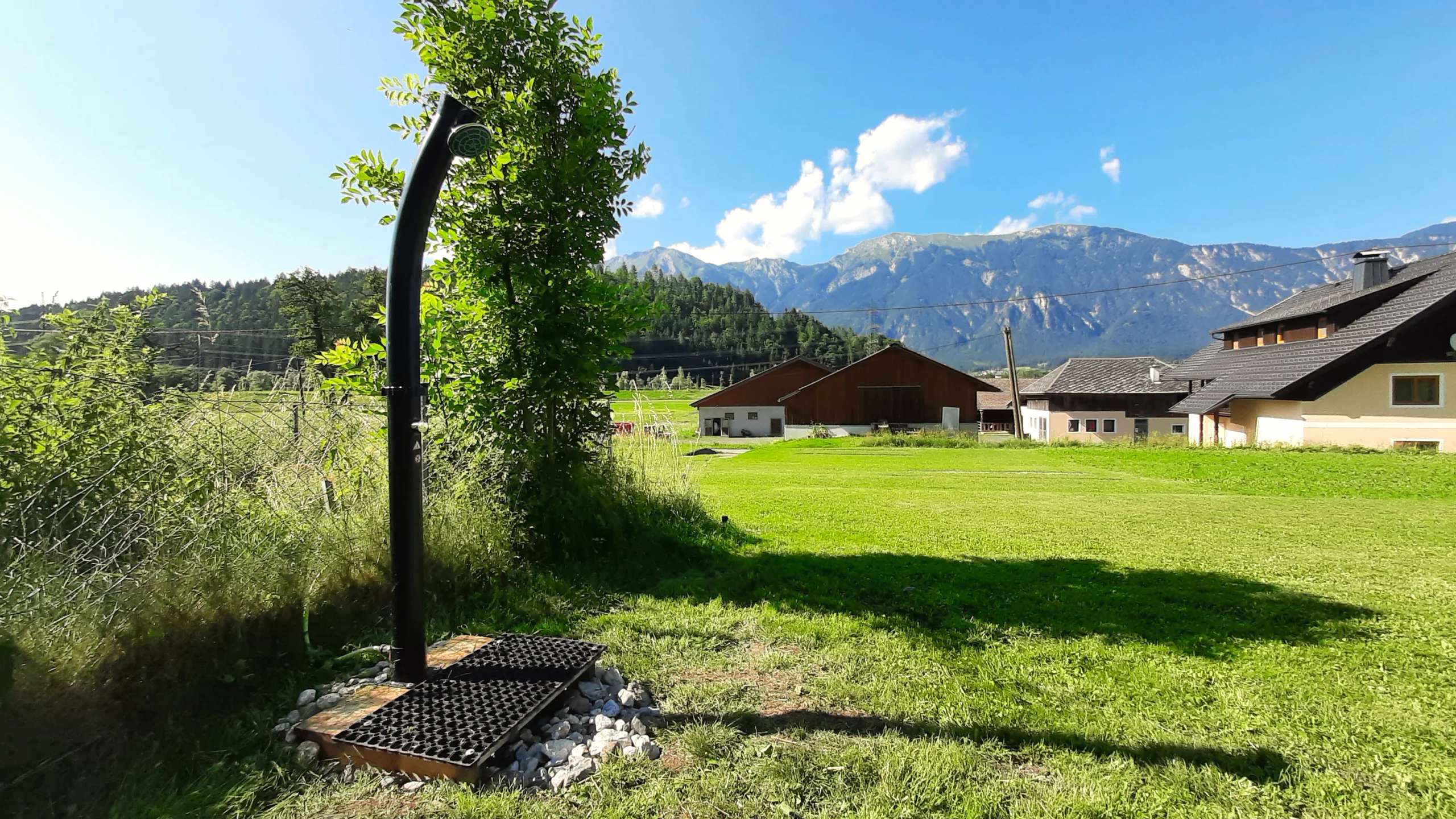 Freistehende Außendusche auf der Wiese, dahinter der Hof und das Bergpanorama