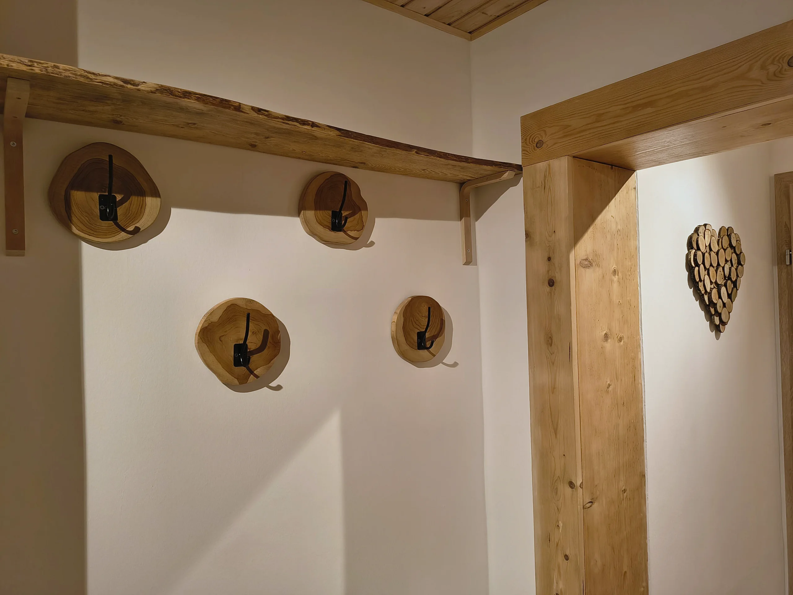 Garderobenhaken auf Holzscheiben an der Wand, darüber ein Balken als Ablage, daneben ein Herz aus Holzstücken