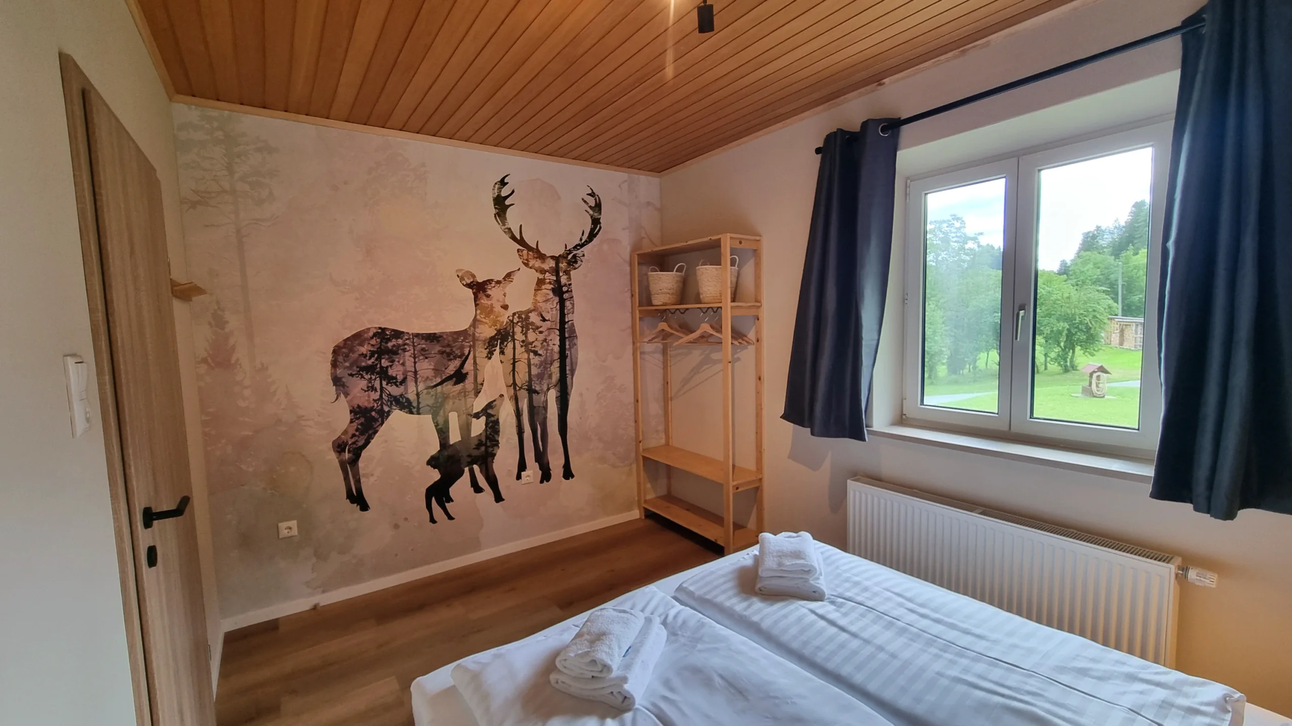 Schlafzimmer der Ferienwohnung 2 mit Hirsch-Wandbild, frischen Handtüchern auf dem Bett und Fenster ins Grüne