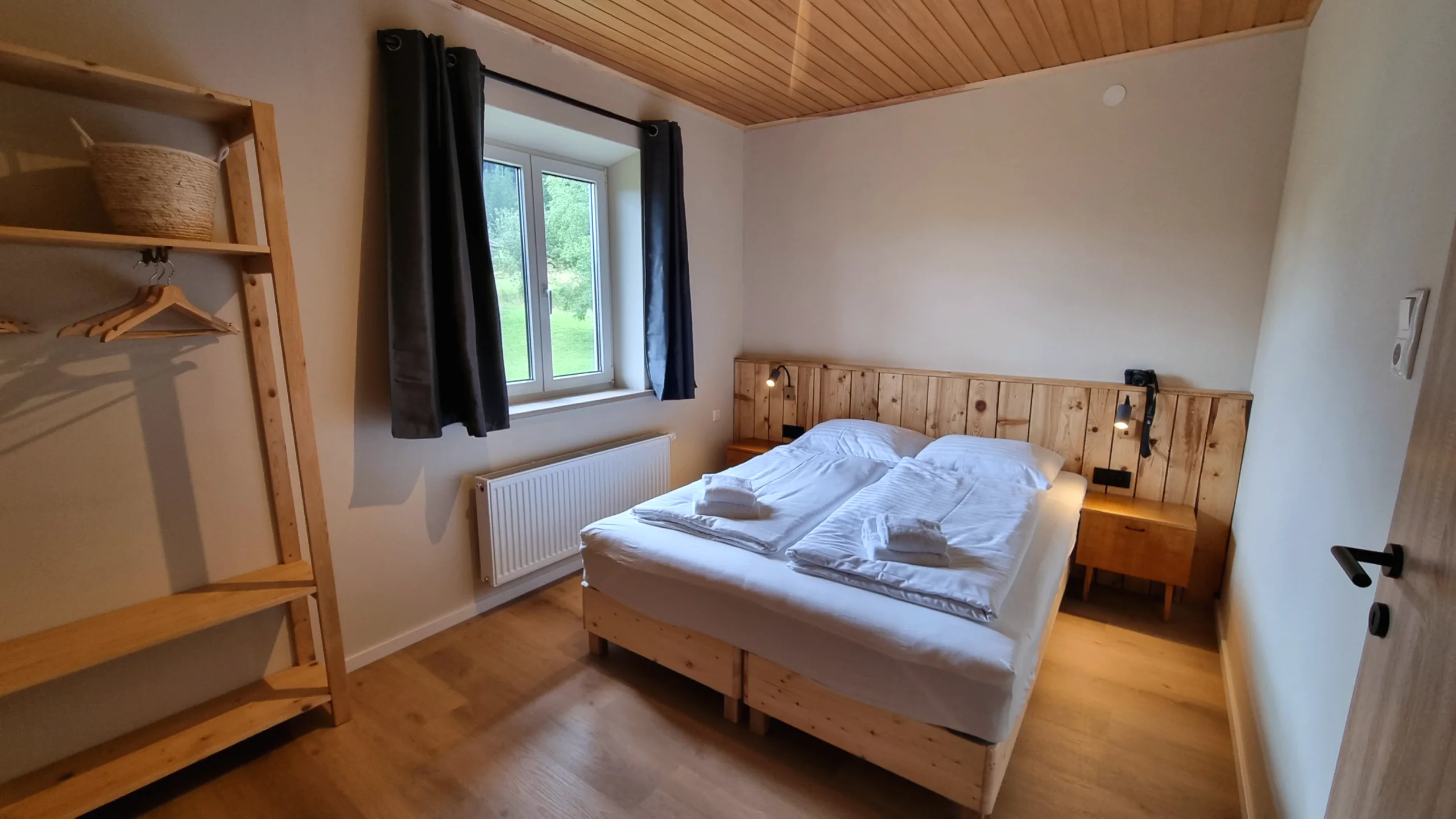 Helles Schlafzimmer mit Holz-Kopfteil, offenem Regal und Fenster ins Grüne