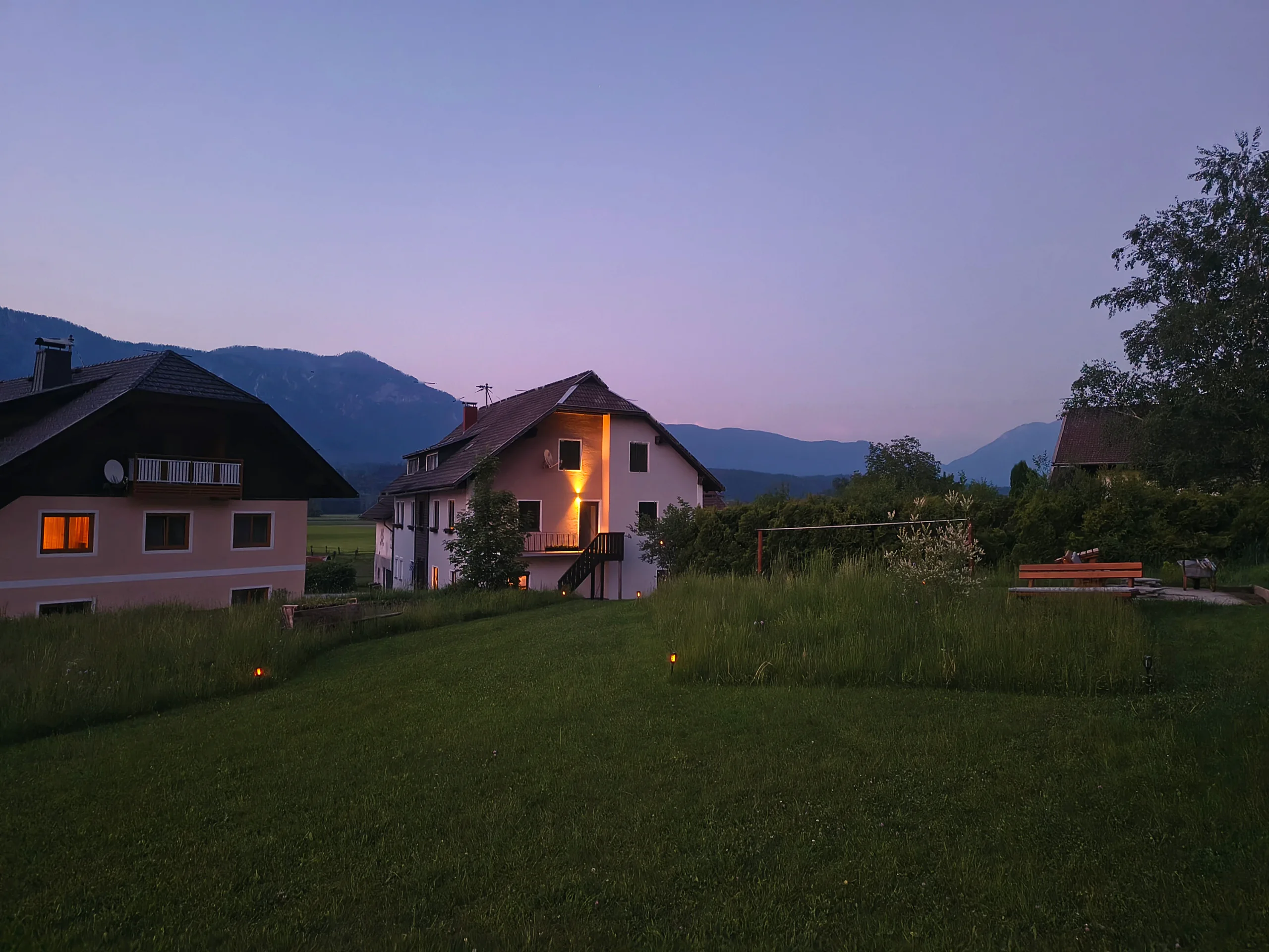 Haus Nampolach im Sommerabendlicht, die Fenster warm erleuchtet, davor die Wiese und dahinter die Gailtaler Berge